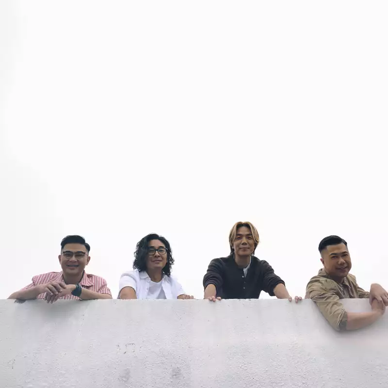 Four members of RubberBand stood behind a wall outside, smiling. The background is grey overcast.
