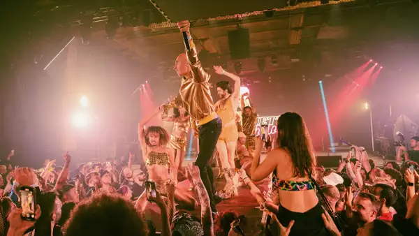 A group of performers pose on a catwalk while a large crowd cheers on and takes photographs. The lighting is pink.