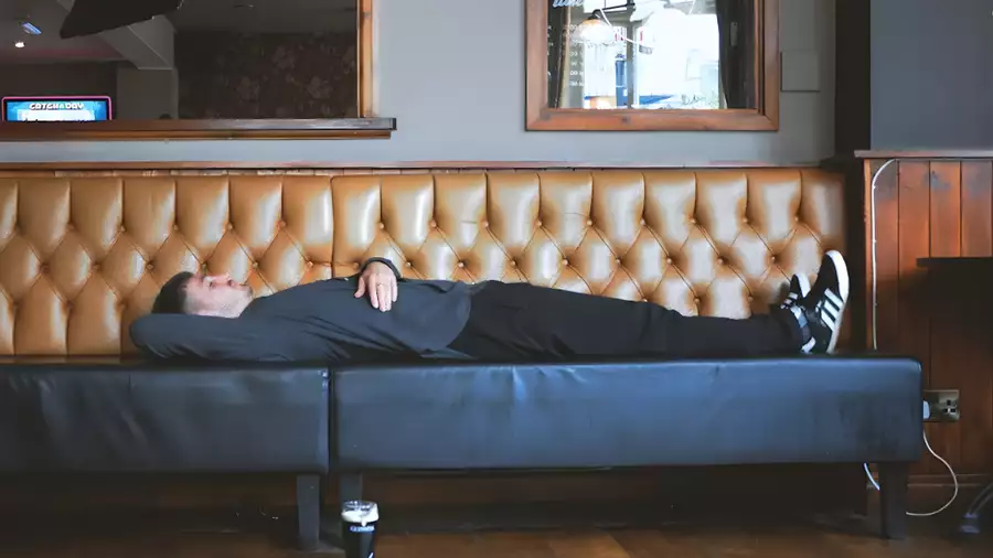 A person lies stretched out on a long tufted leather bench in a dim bar or café interior, resting with one arm under their head and a drink on the floor nearby.