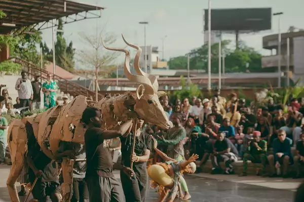 Performers operate a large cardboard antelope puppet in front of a seated outdoor audience.