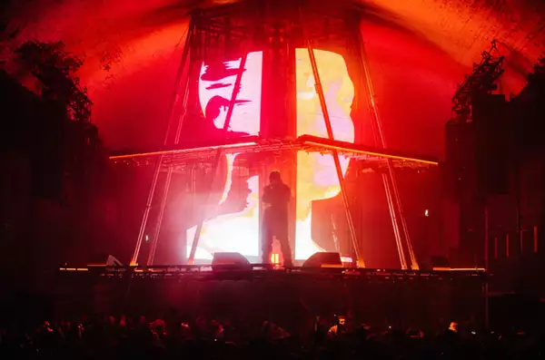 A photograph of Skepta, stood in silhouette on a brightly lit stage. The stage is a custom-built pyramid with a flat top, with a centrepiece screen showing 3D graphics of a human head shown across multiple large LED displays