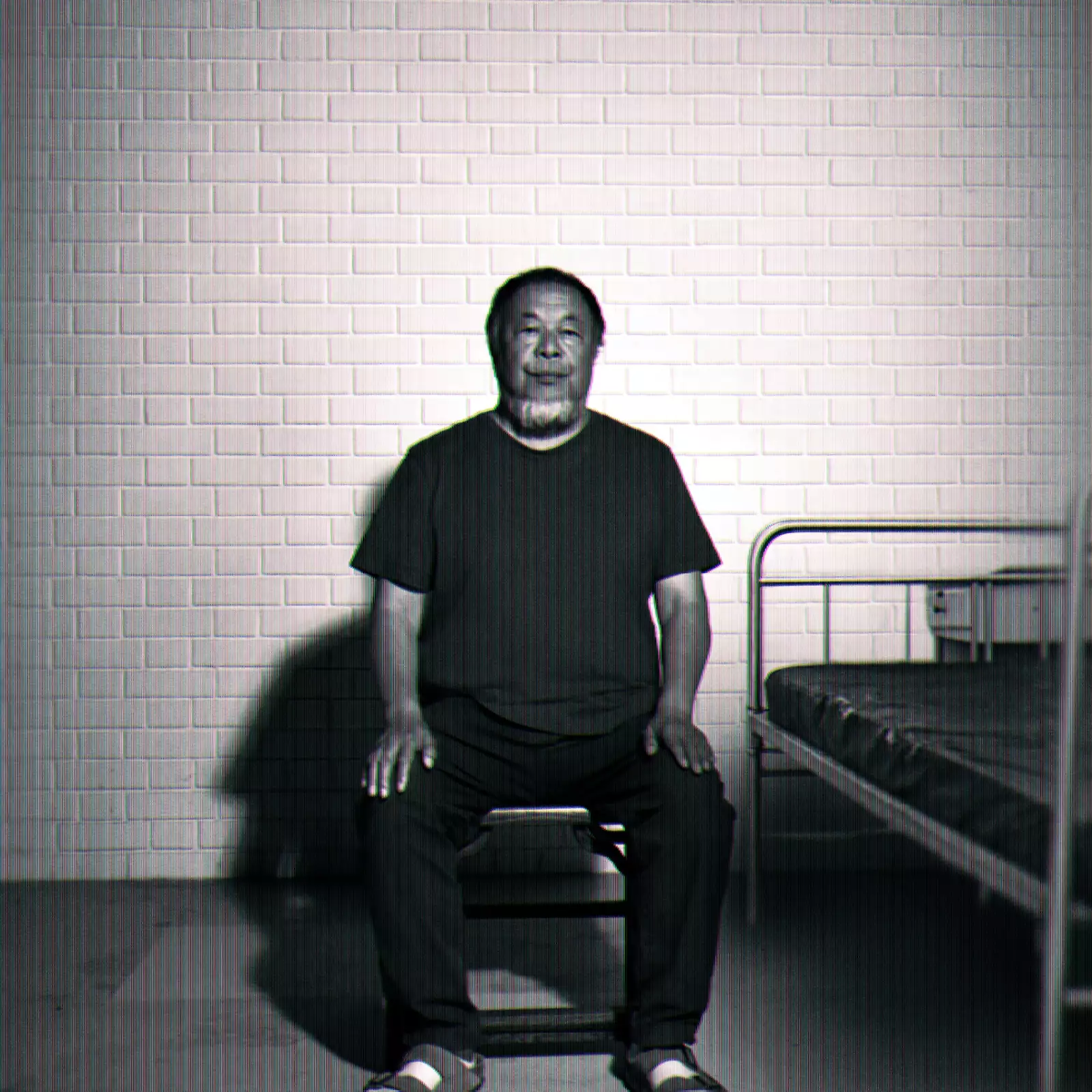 Black-and-white image of Ai Weiwei sitting on a chair beside a simple metal bed against a brick wall.