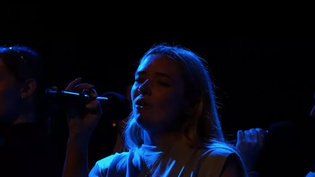 Lizzie Simms performing on stage, singing into a microphone. The photo is dark with a blue hue.