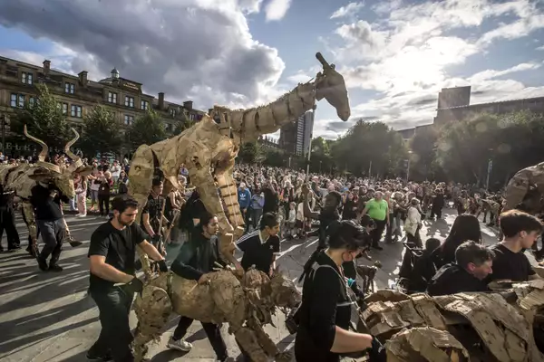 Performers parade life-sized cardboard animal puppets through a city square, watched by a large crowd on a sunny day.
