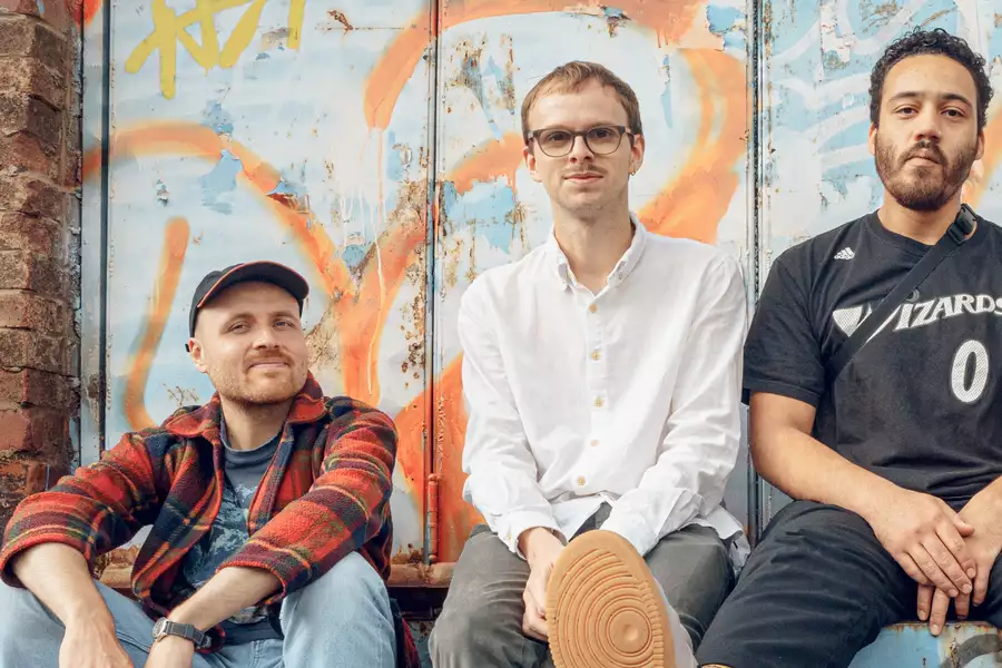 Three men sit casually on a graffitied blue metal ledge in front of a brick wall and colourful, spray-painted backdrop. The man on the left wears a red plaid shirt, light blue jeans, and a cap. The man in the middle, dressed in a white button-up and glasses, has one leg crossed with the sole of his shoe facing the camera. The man on the right wears a black Wizards basketball jersey and rests his hands in his lap. The trio exudes a relaxed, urban vibe.