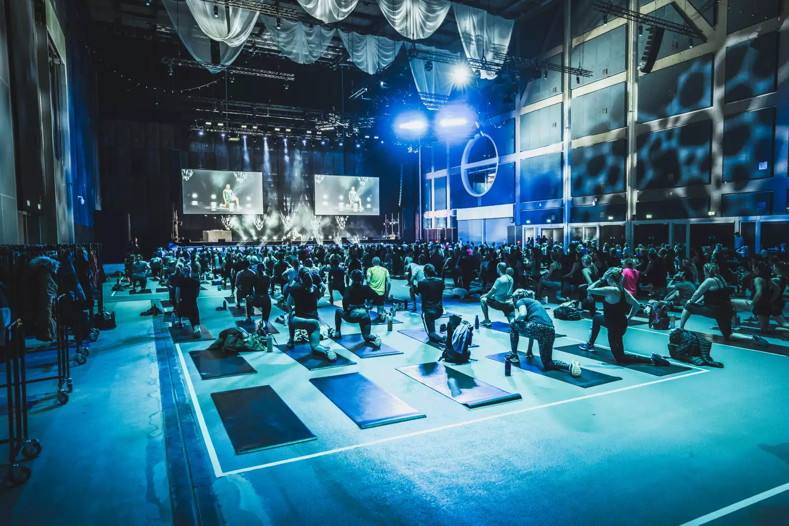 A large yoga session in the Warehouse during MOVE Manchester. The lighting is blue. At the front is a stage with two large screens.