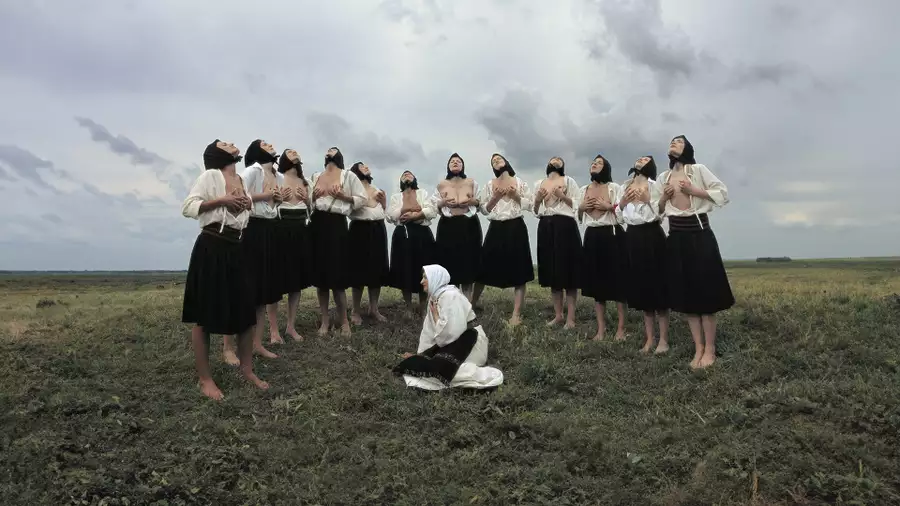 Artist Marina Abramovic kneels on a grassy field in a white robe, surrounded by a group of women stood in a semi-circle in black skirts, white shirts and black headscarves all holding their breasts and looking up at the sky