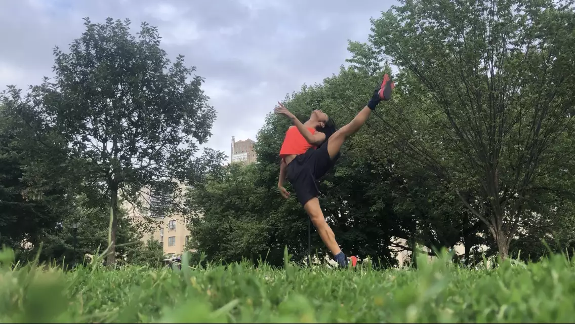 A horizontal photo of ​​Benedict Nguyễn, wearing black shorts, orange tank, and black and orange sneakers, standing on right leg with left leg raised to the side, left arm bent in front of her torso, head tipped back, in a field with green grass and trees, with buildings in distant background.