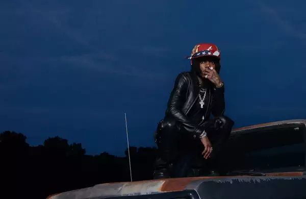 J.I.D wearing a black leather outfit and red hat. He squats on top of an old car at dusk.