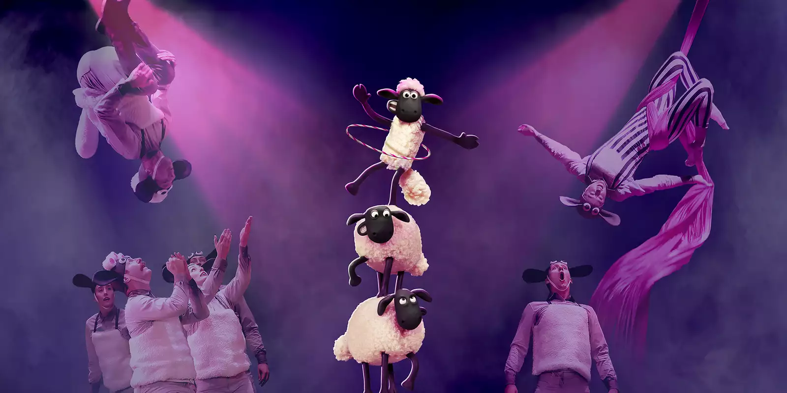 Performers in sheep costumes execute acrobatic stunts and aerial silk acts under dramatic pink lighting. In the center, three animated sheep from "Shaun the Sheep" balance in a tower, with the top one hula-hooping.