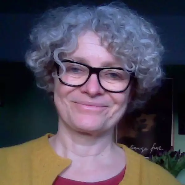 A woman with short curly grey hair smiles at the camera. She wears black rimmed glasses, a yellow cardigan and red t-shirt.