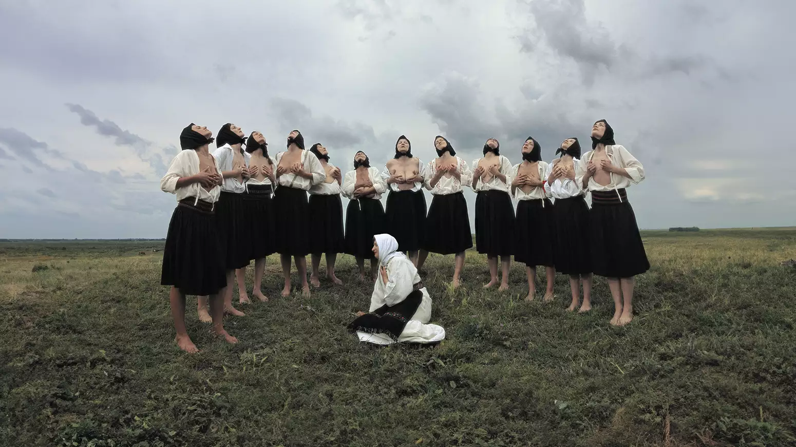 A group of women in traditional Eastern European attire stand barefoot in a grassy field under a dramatic, cloudy sky. Most are wearing black skirts, white blouses, and black headscarves, with some partially exposing their chests while lifting their heads upward in a ritualistic pose. In the foreground, a single woman dressed in white kneels on the ground with a headscarf and embroidered belt, gazing to the side.