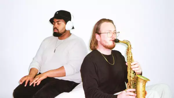Two members of Secret Night Gang sat down against a white background. One has headphones on with their eyes closed. The other plays a saxophone.