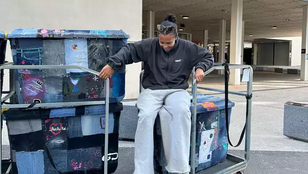 A person in a dark sweatshirt and grey sweatpants sits on a cart next to two large bins covered in colourful, patchwork denim fabric, outdoors near Aviva Studios.