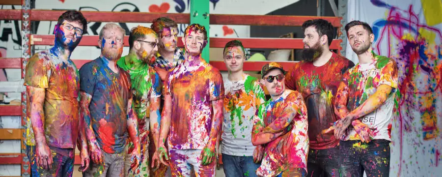 Nine members of Young Pilgrims covered in colourful paint, posing in an artistic setting.