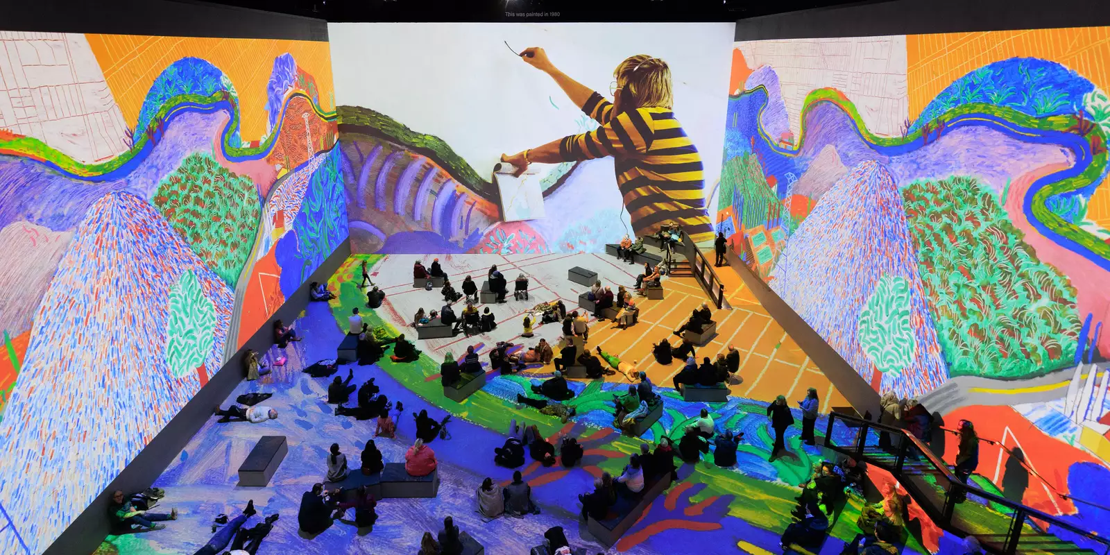 Photograph of David Hockney: Bigger & Closer (not smaller & further away) at Lightroom taken from above. It shows people sitting and lying on the floor, immersed inside Hockney's artworks.