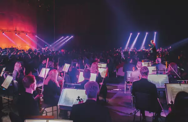 The BBC Philharmonic orchestra performing in the Warehouse. The lighting is pink and purple.  The audience are stood in the middle of the orchestra.