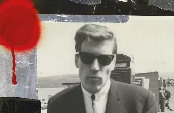 A black-and-white photo of a man wearing sunglasses and a suit, with a blurred background of a seaside scene. There is a bright red circle of paint on the photograph.