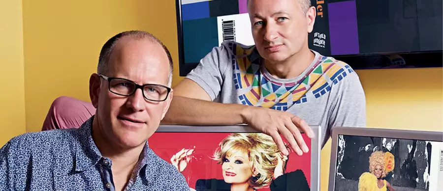Two people posing with framed photos of performers against a yellow wall