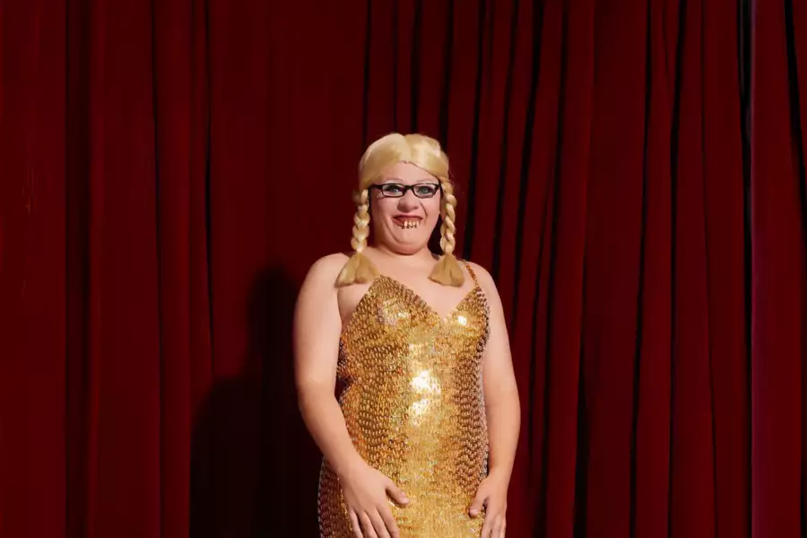 Bailey J Mills in a gold glittery dress wearing a plaited blonde wig, stood infront of red curtains