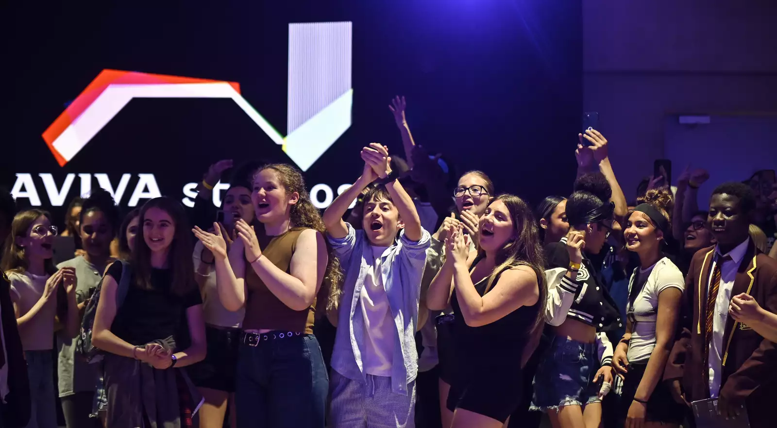 A group of children clapping and cheering in the Social – with a screen behind them that says 'Aviva Studios'