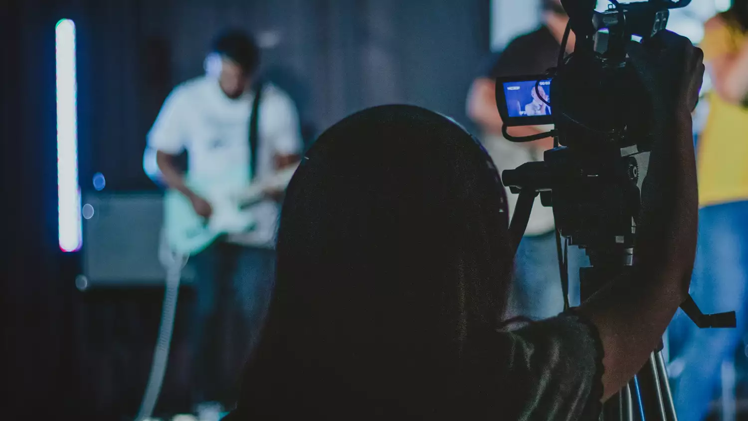 A person wearing headphones behind a video camera films a band playing onstage