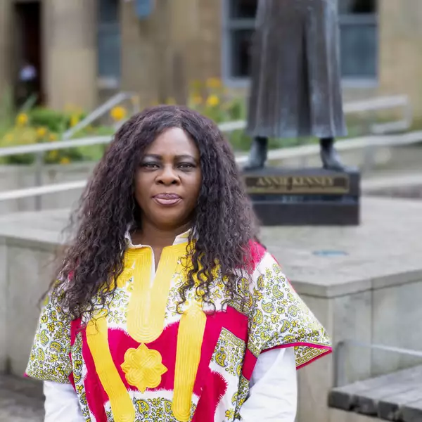 Headshot of Chantelle Tindall wearing a red and yellow patterned top. They are stood outside in front of a statue of the suffragette Annie Kenney.