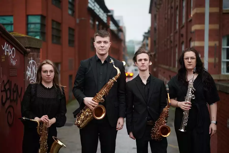 The Akiko Sax Quarte stand amongst redbrick buildings in all black clothing holding their instruments