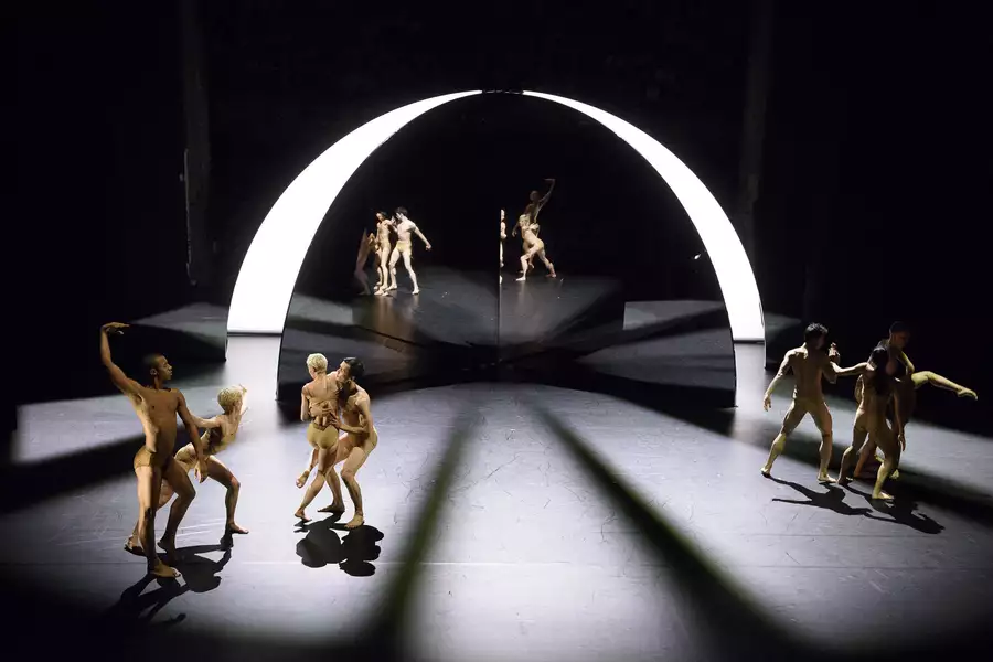 Seven dancers are posed in front of an illuminated archway. A mirror behind them makes it look as though there are more dancers present.
