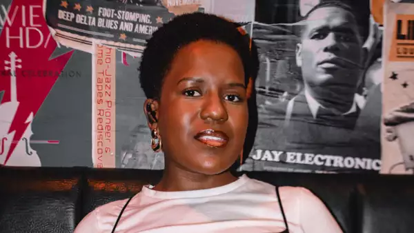 Rumbi Tauro wearing a white top and gold earrings, stood in front of a collaged wall