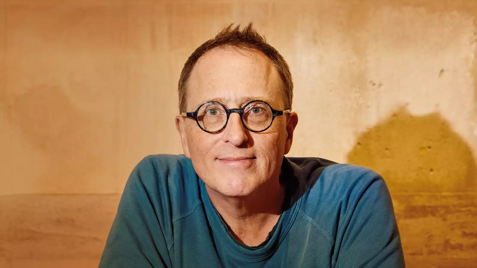 Jon Ronson smiling in front of a wall, wearing a blue t-shirt and glasses