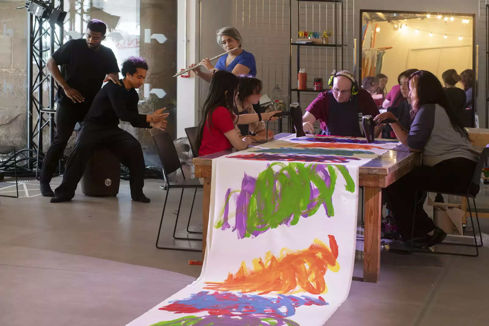 A photo of artist Michael Beard painting on a long roll of paper while a flautist from Manchester Camerata plays nearby and dancers from Company Chameleon perform to the music