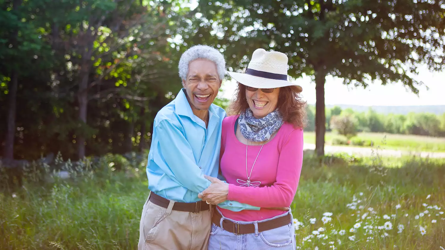 Glenn and Elizabeth Copeland smiling, laughing and hugging outside in a field. Glenn is wearing a bright blue shirt. Elizabeth is wearing a bright pink top.