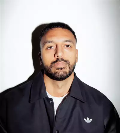 Ayman Chaudhry wearing a white t-shirt and zipped up black adidas jacket, on a white background