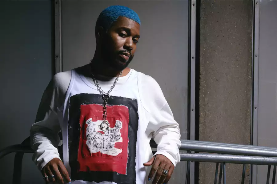 Khalid with blue hair wearing a white shirt and chain necklace, standing against a wall.