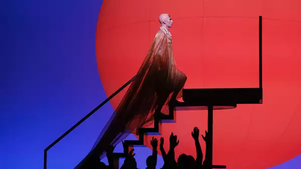 An opera stage, showing one person climbing steps in an opulent dress with others raising their hands towards them. The background is red and blue.