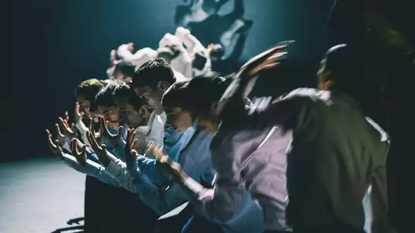 A group of dancers stood in a line with their arms extended forward, moving in blurred motion. The lighting is dim, casting dramatic shadows.