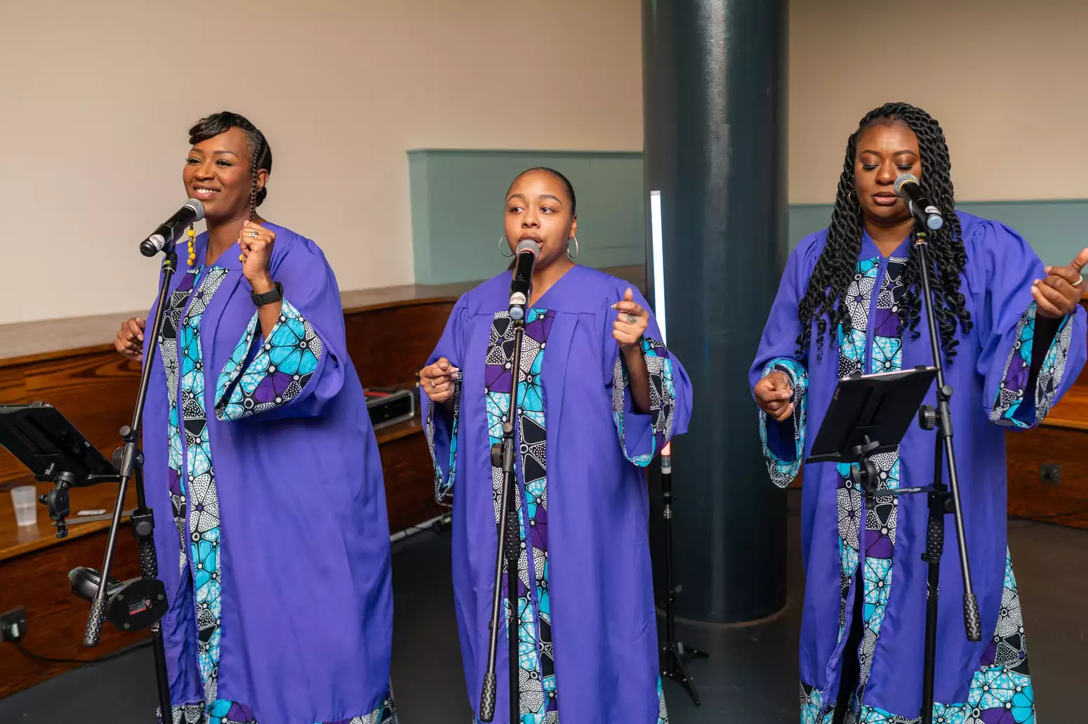 Three women are singing into microphones in the Lower Hall Foyer at Aviva Studios. They are wearing purple traditional dress.