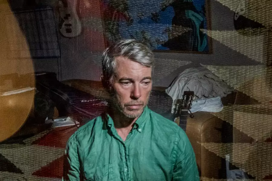 Bill Callahan wearing a green shirt sitting indoors, overlaid with a double exposure of a guitar and patterned rug.