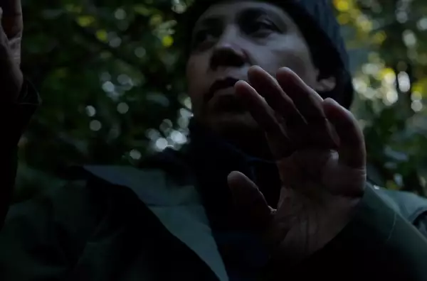 A person stands against foliage with their hands raised. They wear a beanie hat and green waterproof jacket.