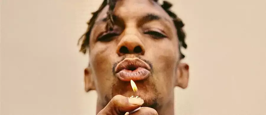 Tricky holds a lighter flame near his mouth, on a beige background.