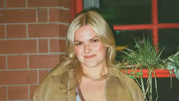 Blythe smiling and wearing a brown jacket. They are stood in front of a red window with plants on the window sill.