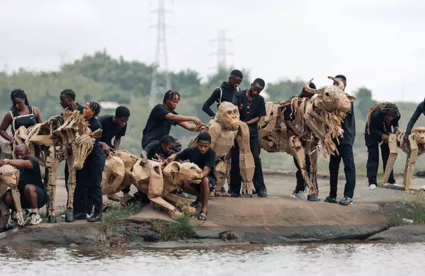 A herd of cardboard animal puppets being operated by people in black clothes by the side of a river