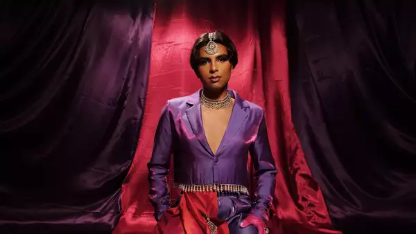 Jason Patel wears a purple and pink suit, against a backdrop of deep purple and pink draped fabric, looking confidently at the camera.