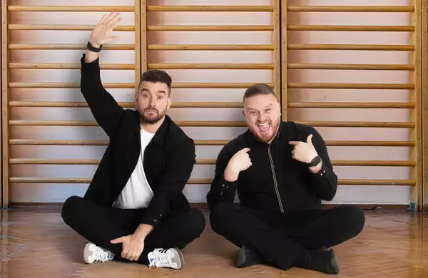 The Two Mr Ps sitting on the floor in front of wooden wall bars, one raising his hand and the other pointing at himself.