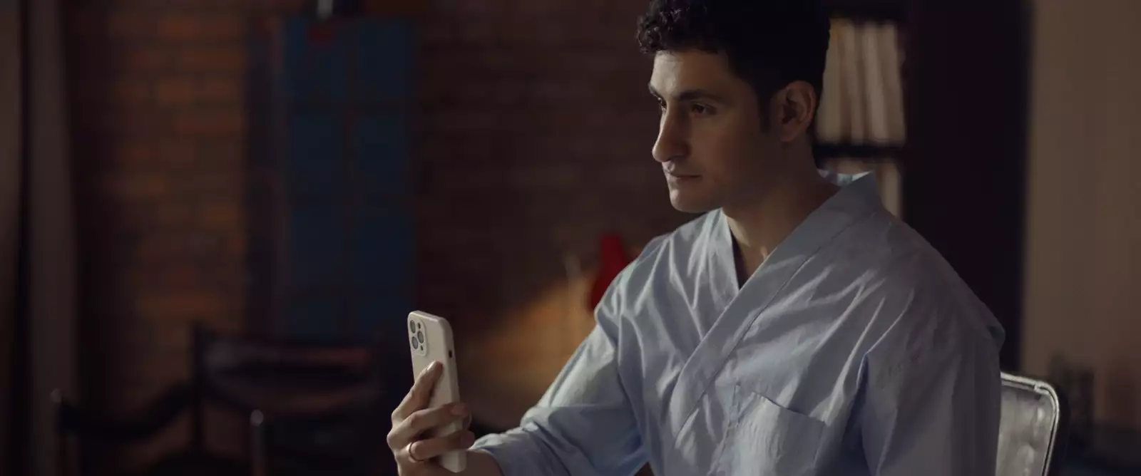 Still from In Camera. Amir El Masry is holding their phone up as if filming something off-screen. They are wearing a light blue robe.