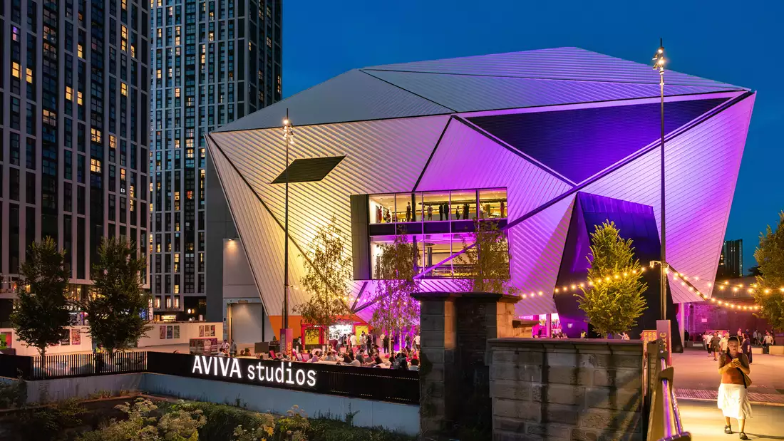 A photo of the venue Aviva Studios lit up with purple lights with crowds enjoying Festival Square by the river
