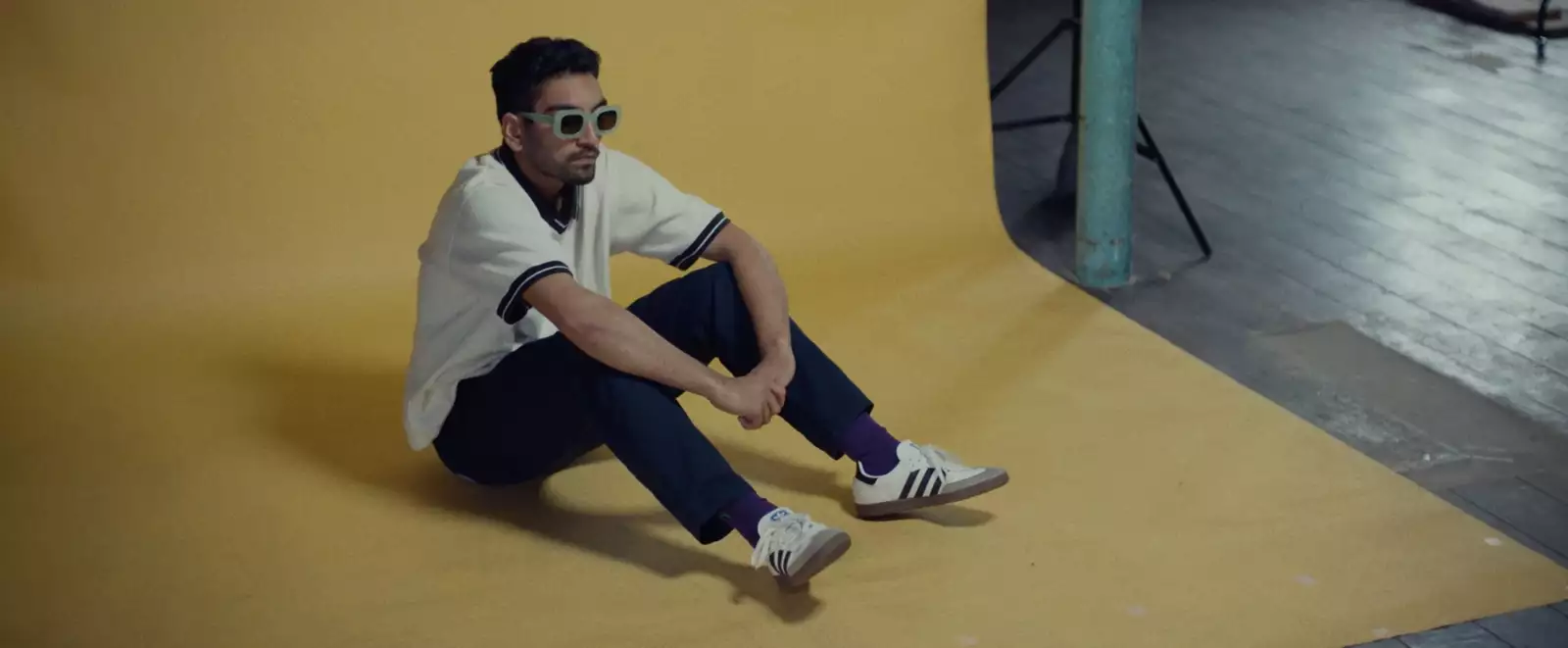 Still from In Camera. Nabhaan Rizwan is wearing sunglasses and trainers, sat against a yellow backdrop on set.