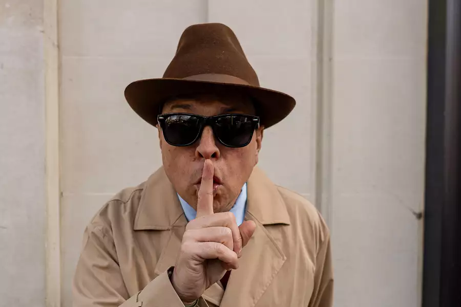 Dom Joly wearing a trench coat, fedora, and sunglasses gestures 'shush' with his finger to his lips."