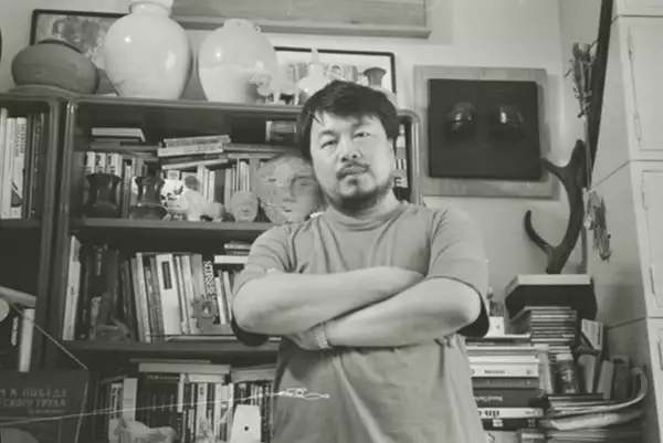 Black and white photograph of Ai Weiwei stood in front of a bookshelf